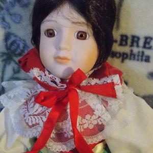 May include: A porcelain doll with dark brown hair, wearing a red and white lace collar with a red bow, and a green and white dress.