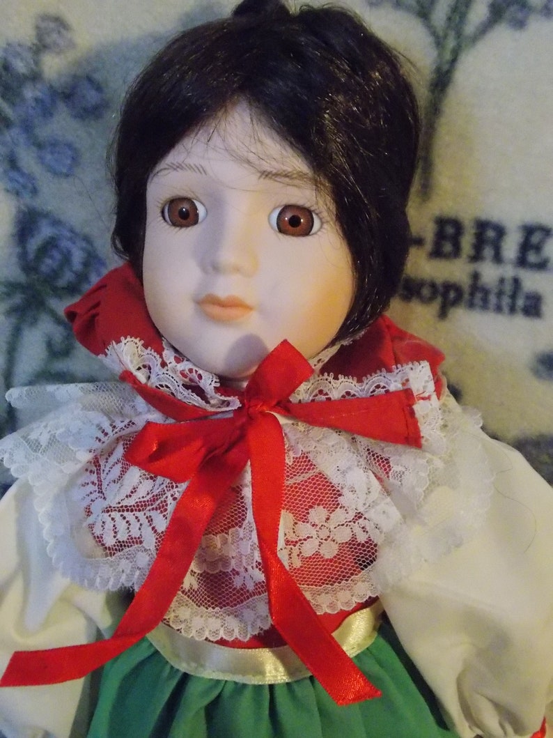 May include: A porcelain doll with dark brown hair, wearing a red and white lace collar with a red bow, and a green and white dress.