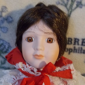 May include: A porcelain doll with brown hair, brown eyes, and a red and white dress. The doll is wearing a white lace collar and a red bow.