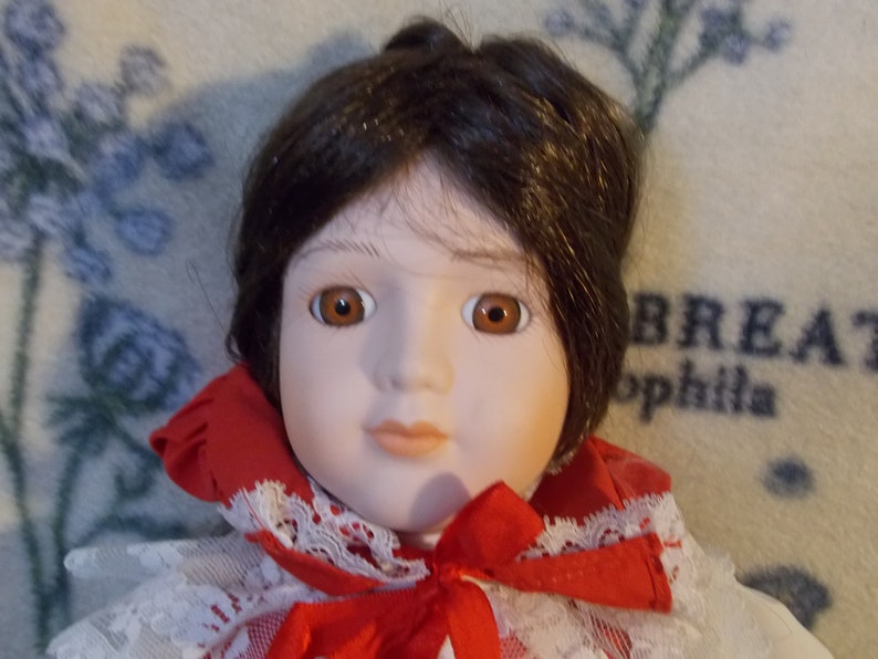 May include: A porcelain doll with brown hair, brown eyes, and a red and white dress. The doll is wearing a white lace collar and a red bow.