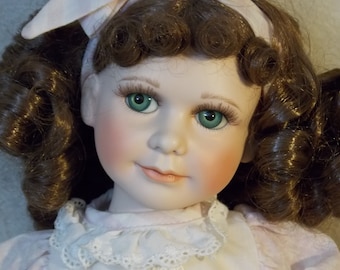 May include: A porcelain doll with brown curly hair, wearing a white dress with lace trim and a pink headband. The doll has green eyes and a painted smile.