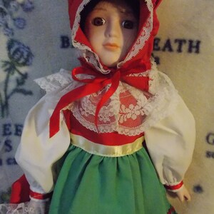 May include: A porcelain doll wearing a red and white dress with a green skirt and a red bonnet. The doll has brown hair and blue eyes.