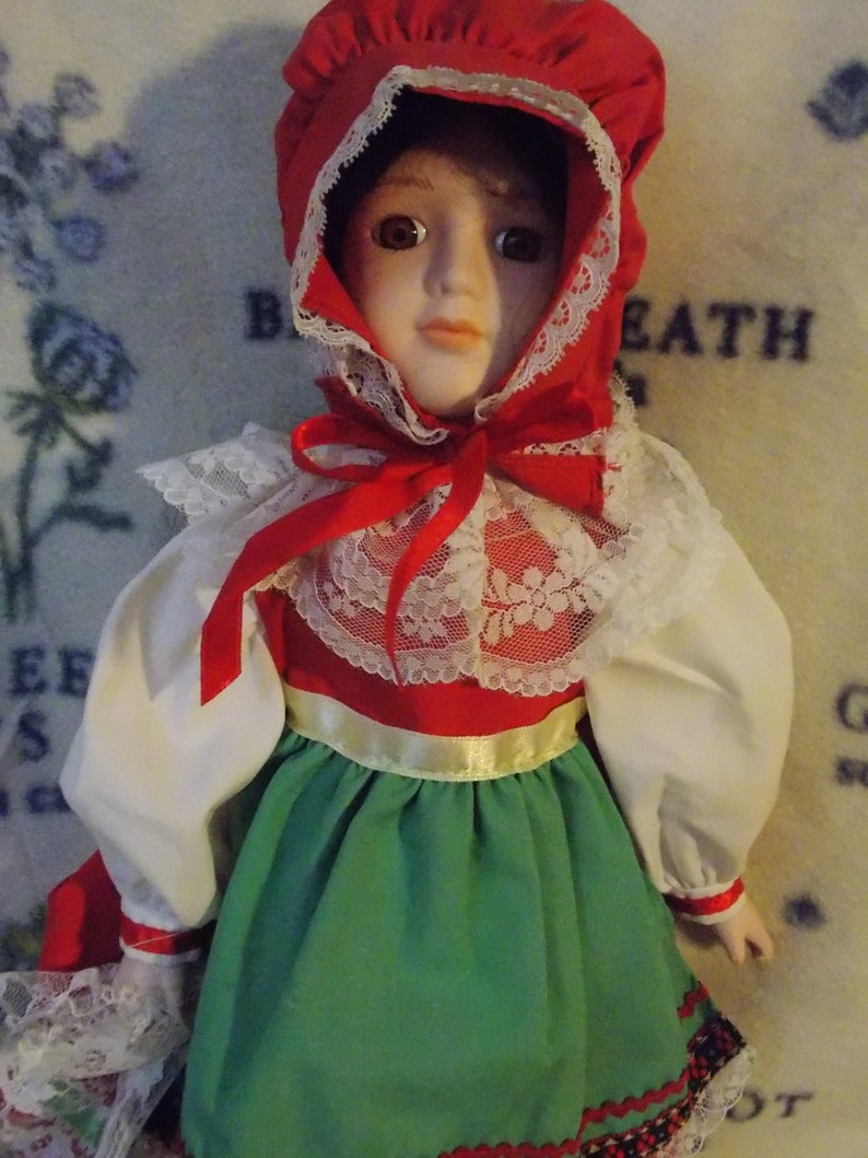 May include: A porcelain doll wearing a red and white dress with a green skirt and a red bonnet. The doll has brown hair and blue eyes.