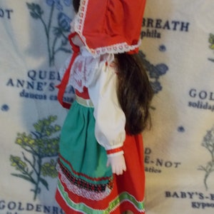 May include: A porcelain doll wearing a red and white dress with a green skirt and a red bonnet. The doll has brown hair and is wearing white shoes.