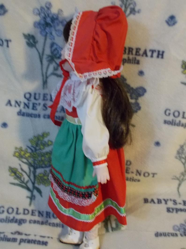 May include: A porcelain doll wearing a red and white dress with a green skirt and a red bonnet. The doll has brown hair and is wearing white shoes.