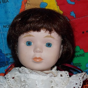 Könnte beinhalten: Eine Porzellanpuppe mit braunen Haaren und blauen Augen, die ein weißes Spitzenkleid mit einem roten Band trägt. Die Puppe sitzt auf einer Weltkarte.