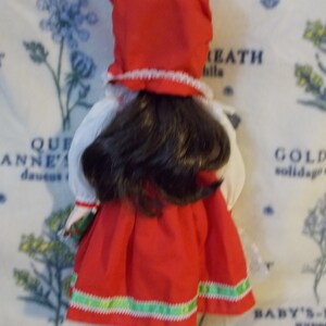 May include: A porcelain doll wearing a red dress with green trim and a red hat with white trim. The doll has long brown hair and is holding a green package.