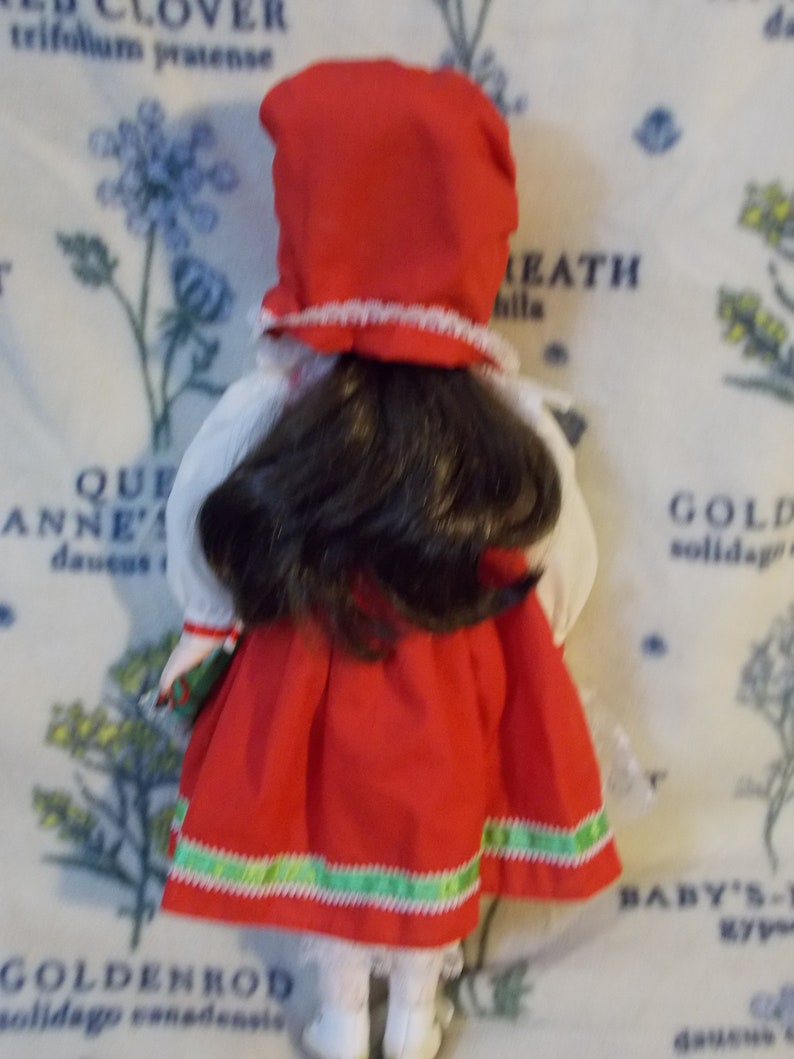 May include: A porcelain doll wearing a red dress with green trim and a red hat with white trim. The doll has long brown hair and is holding a green package.