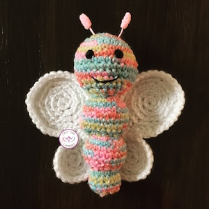 Könnte beinhalten: Ein farbenfrohes gehäkeltes Schmetterlings-Spielzeug mit weißen Flügeln und einem lächelnden Gesicht. Der Schmetterling ist aus verschiedenen Farben hergestellt, darunter Pink, Blau, Gelb und Grün.