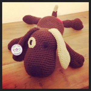 Puede incluir: Un perro de crochet marrón con orejas beige y nariz roja. El perro está acostado sobre una superficie de madera. El perro tiene una etiqueta que dice "First Twin Company".