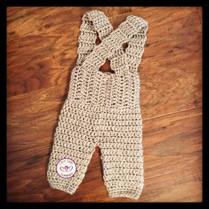 May include: A crocheted baby romper in a light brown color with criss-cross straps. The romper has a small logo on the leg that says "First Year Company".