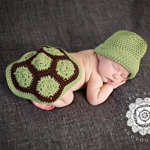 May include: A newborn baby wearing a green and brown crocheted turtle costume, sleeping on a gray textured surface. The baby is wearing a green crocheted hat.