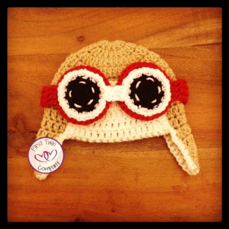 May include: A crocheted baby hat with a brown, white, and red color scheme. The hat features a pair of goggles with black lenses and white stitching. The hat has a flap that covers the ears. The hat is made by First Twin Company.