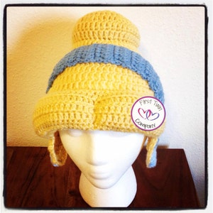May include: A yellow and blue crocheted hat with a bun on top. The hat has ear flaps and a small tag that says "First Twin Company" with two pink hearts.