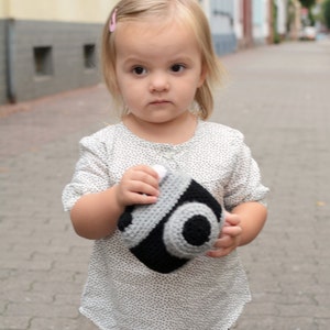 May include: A young girl with blonde hair is holding a black and white crocheted camera toy. She is wearing a white and grey polka dot shirt and grey pants.