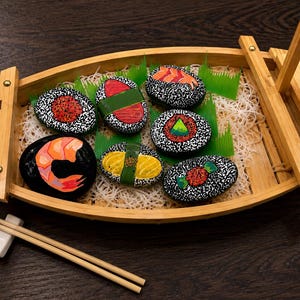 May include: A wooden sushi boat serving platter containing six hand-painted river stones designed to resemble various sushi pieces, including nigiri and maki rolls, resting on shredded white paper and green plastic grass, with wooden chopsticks.