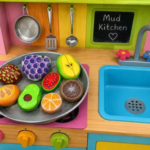May include: A wooden toy kitchen with a blue sink, a faucet with flower-shaped handles, and a chalkboard labeled Mud Kitchen. A metal pan sits on the stove containing painted rock food items including grapes, watermelon, orange slices, and avocado.