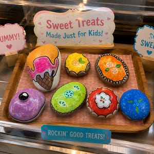 May include: A wooden tray displays six hand-painted rocks featuring colorful designs of ice cream, citrus slices, a donut, and berries. Decorative signs with the text Sweet Treats Made Just for Kids, YUMMY, SO SWEET, and ROCKIN' GOOD TREATS are included.