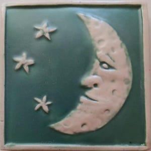 May include: A square ceramic tile featuring a raised, cream-colored crescent moon with a face and three small, five-pointed stars against a smooth, matte teal-green background. The tile has a raised border edge in a matching cream color.