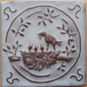May include: Square ceramic tile with a raised relief design. The design features a bird feeding its young in a nest, surrounded by a circular frame and decorative flourishes. The tile is light gray with brown accents.