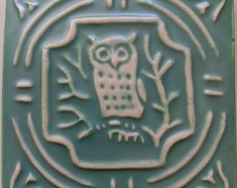May include: A square ceramic tile featuring a raised relief design in a sage green and off-white color scheme. The center displays an owl perched on a branch within a decorative border, surrounded by geometric patterns and stylized corner accents.
