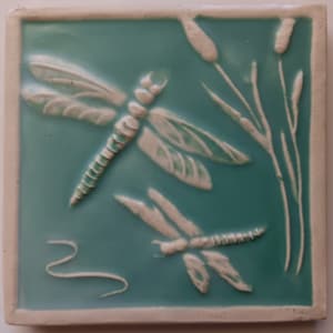 May include: A ceramic tile with a blue-green background and two white dragonfly designs. The dragonflies are in relief and have delicate wings. The tile also features white stylized reeds.