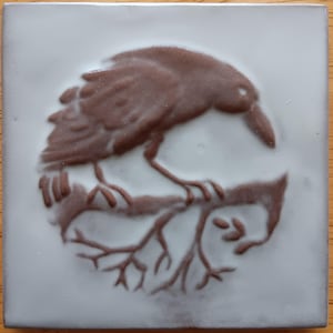 May include: Square ceramic tile with a brown relief image of a crow perched on a branch. The crow is facing right, with its beak pointed down. The tile has a light gray background.