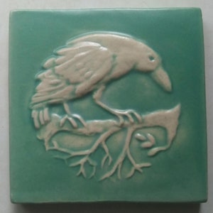 May include: A white ceramic tile with a raised design of a crow perched on a branch. The tile is a light teal color.