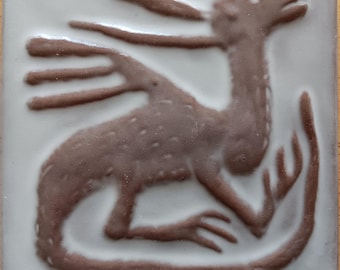 May include: A brown ceramic tile with a raised design of a stylized dragon with wings and a long tail. The dragon is facing right and has a spotted body.