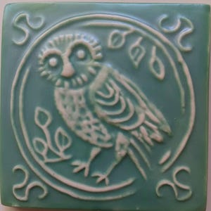 May include: A square ceramic tile in a muted sage green color featuring a raised, light-colored relief design of an owl centered within a circular border. The owl is surrounded by stylized leaf motifs, and each corner of the tile displays a swirl pattern.
