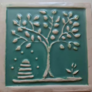 May include: A square ceramic tile with a teal glazed surface featuring a raised white relief design. The design depicts a central tree with numerous leaves, a stacked stone cairn to the left, and a small sprout on the right side.