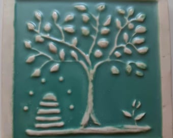 May include: A square ceramic tile with a teal glazed surface featuring a raised white relief design. The design depicts a central tree with numerous leaves, a stacked stone cairn to the left, and a small sprout on the right side.