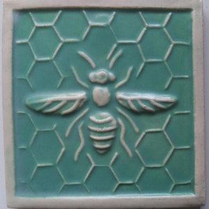 May include: A ceramic tile with a white bee design on a teal blue background. The bee is centered on a honeycomb pattern.