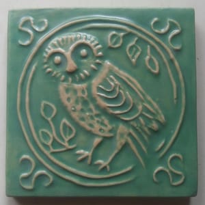 May include: A green ceramic tile with a white owl design. The owl is surrounded by a circle with a vine design. The tile has a raised design and a glossy finish.