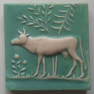 May include: A white ceramic tile with a raised image of a cow standing in a field. The cow is white with a brown head and horns. The field is green with a small tree and a flower.