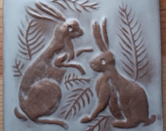 May include: A ceramic tile with a white background and two brown rabbits in relief. The rabbits are surrounded by stylized brown leaves.