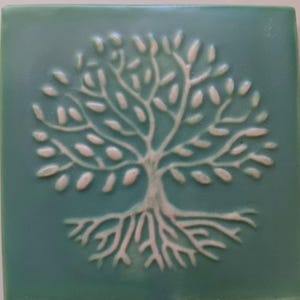 May include: A square ceramic tile with a glossy sage green finish featuring a raised, embossed white design of a tree with visible roots and branches. The tree is centered on the tile, showcasing a smooth texture and a distinct relief pattern.