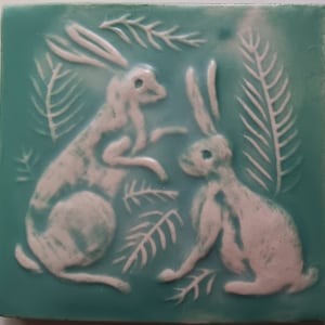 May include: Two white ceramic rabbits are depicted in relief on a turquoise blue square tile. The rabbits are surrounded by stylized white fern leaves.