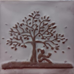May include: A square ceramic tile with a glossy light gray finish featuring a raised, dark brown embossed design of a large tree with a person sitting underneath it, reading a book. The relief texture creates a tactile surface on the tile.