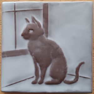 May include: A square ceramic tile featuring a raised, brown-toned relief of a cat sitting in profile. The cat is positioned in front of a simple window frame design. The background of the tile is a smooth, off-white glaze with a matte finish.