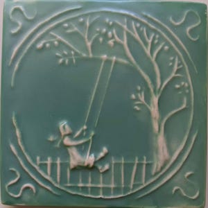 May include: A square ceramic tile in a muted sage green color featuring a raised, light-colored relief design. The central circular motif depicts a figure on a swing suspended from a tree branch, set against a background of a simple picket fence.