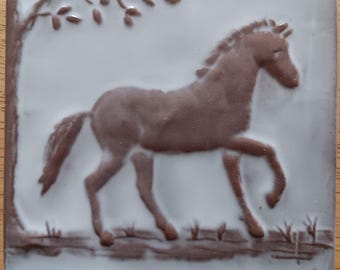 May include: A square ceramic tile featuring a brown horse in relief against a light gray background. The horse is depicted in motion, with a tree branch and foliage in the upper left corner. Grass and a fence are at the bottom.