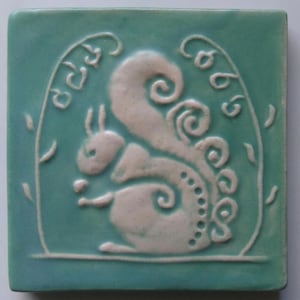 May include: A teal blue ceramic tile with a white raised design of a squirrel with a curly tail. The tile has the word "cheer" written in cursive on the top left corner.
