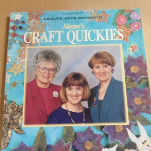Può includere: Una copertina di libro con tre donne sorridenti che tengono in mano materiali per l'artigianato. Il titolo del libro è "Aleene's Craft Quickies" e il sottotitolo è "Leisure Arts Presents". La copertina è decorata con fiori colorati, stelle e altri oggetti per l'artigianato.