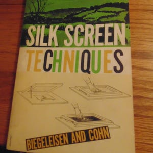 May include: A vintage book titled "Silk Screen Techniques" by Biegeleisen and Cohn. The cover features a green landscape with a white tree and illustrations of silk screen printing techniques.
