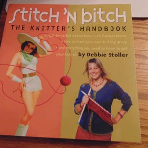 Puede incluir: Un libro titulado "Stitch 'N Bitch: The Knitter's Handbook" de Debbie Stoller. La portada del libro presenta a una mujer con un vestido blanco y un sombrero rojo, sosteniendo una aguja de tejer. La portada también presenta a una mujer con un suéter azul, sosteniendo una aguja de tejer y lana.