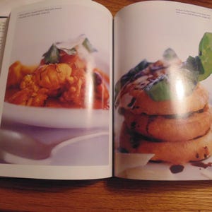May include: Two-page spread from a cookbook featuring two food dishes. The left page shows a white bowl with a reddish-orange dish. The right page displays a stack of golden-brown food items with a dark sauce and green garnish.
