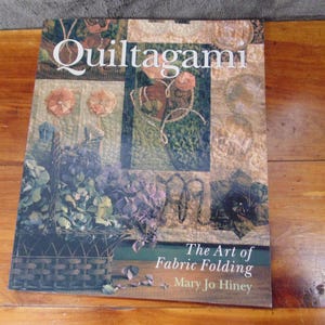 May include: Book cover with the title "Quiltagami: The Art of Fabric Folding" by Mary Jo Hiney. The cover features a colorful quilt with floral designs and a basket of flowers.