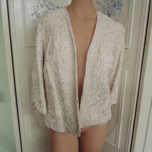May include: A white beaded cardigan with a zipper closure. The cardigan is made of a soft, textured fabric and has a relaxed fit.