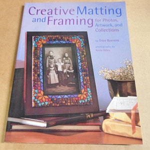May include: A book titled "Creative Matting and Framing for Photos, Artwork, and Collections" by Trice Boerens with photography by Kevin Dilley. The book cover features a close-up of a framed photo with a mosaic frame.
